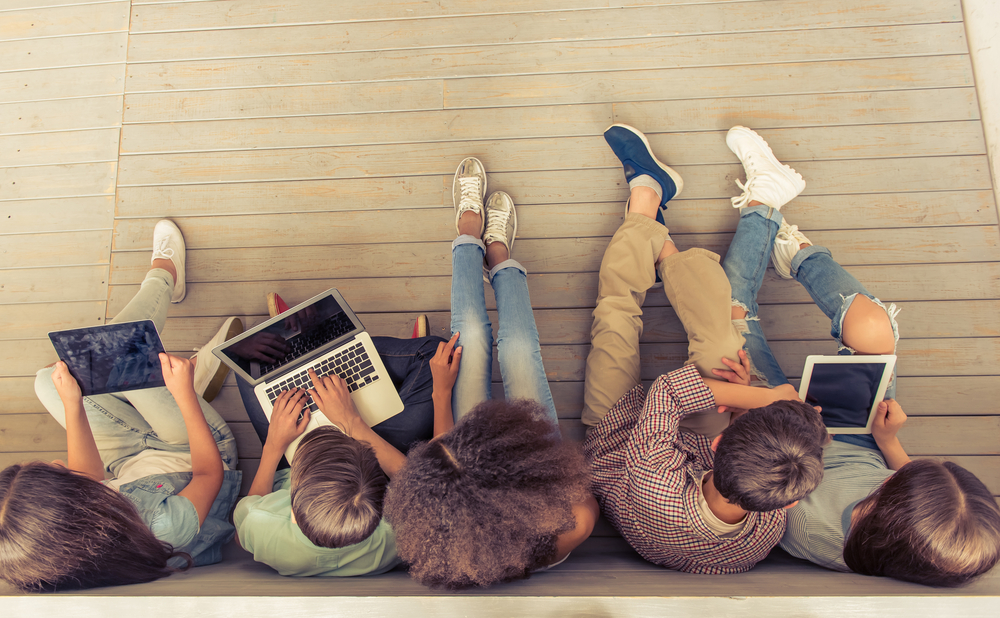 Top view of group of teenage boys and girls using gadgets while sitting in row on wooden floor