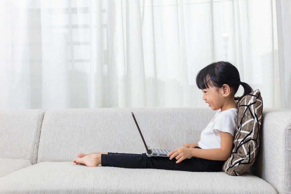 Happy Asian Chinese little girl lying on the sofa with laptop in the living room at home.